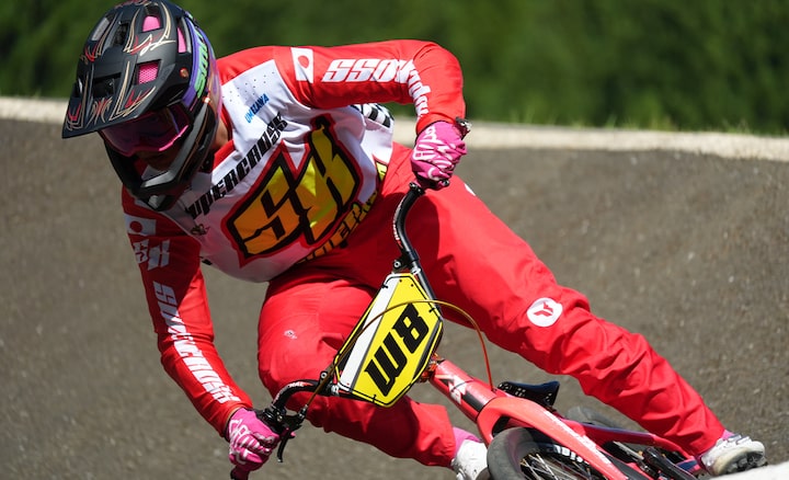 Ein BMX-Fahrer in leuchtend roter Rennmontur lehnt sich während eines Highspeed-Rennens auf einer Schotterpiste tief in eine Kurve.